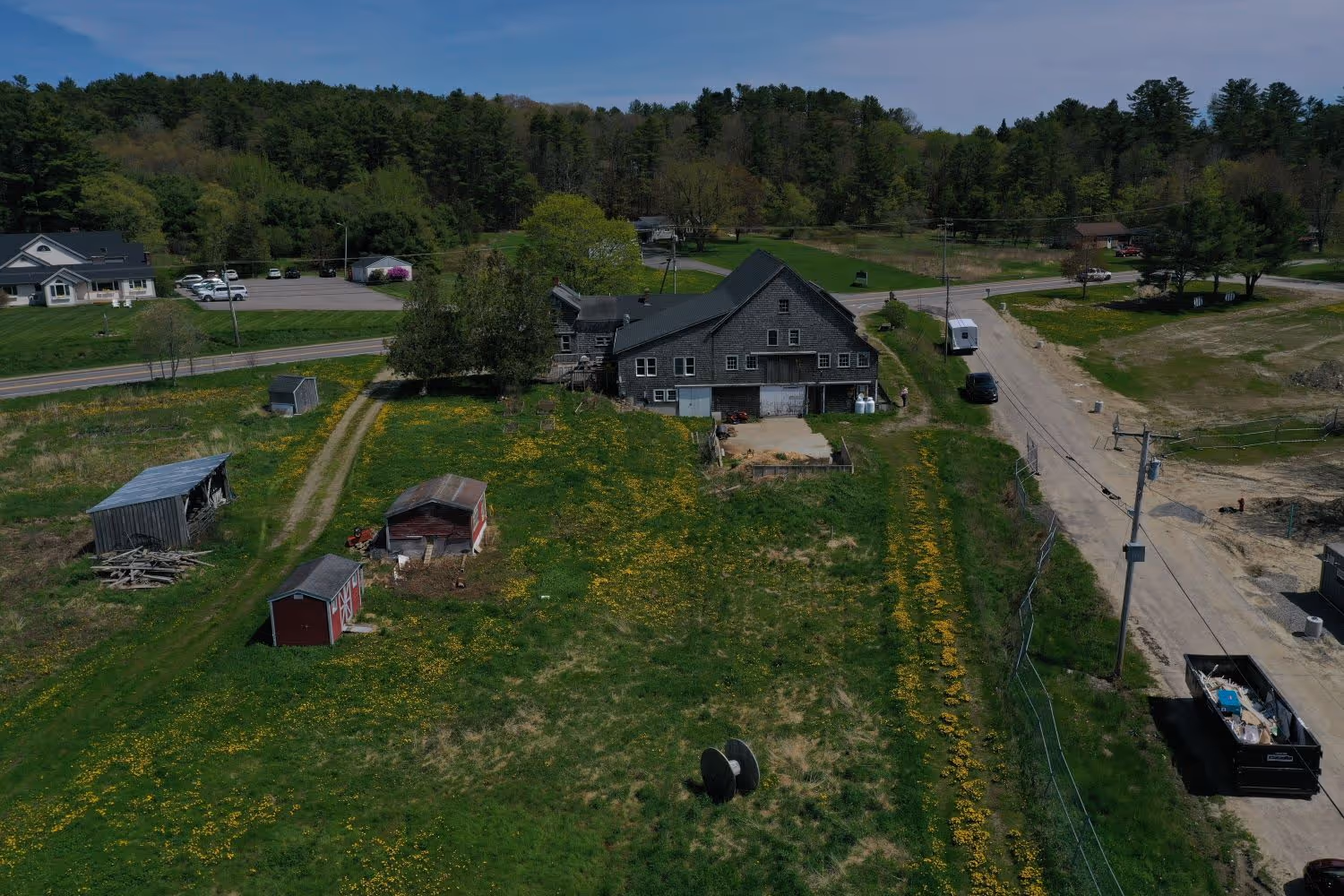 Aerial view of rural farmstead with barns, yellow wildflowers, and surrounding forest