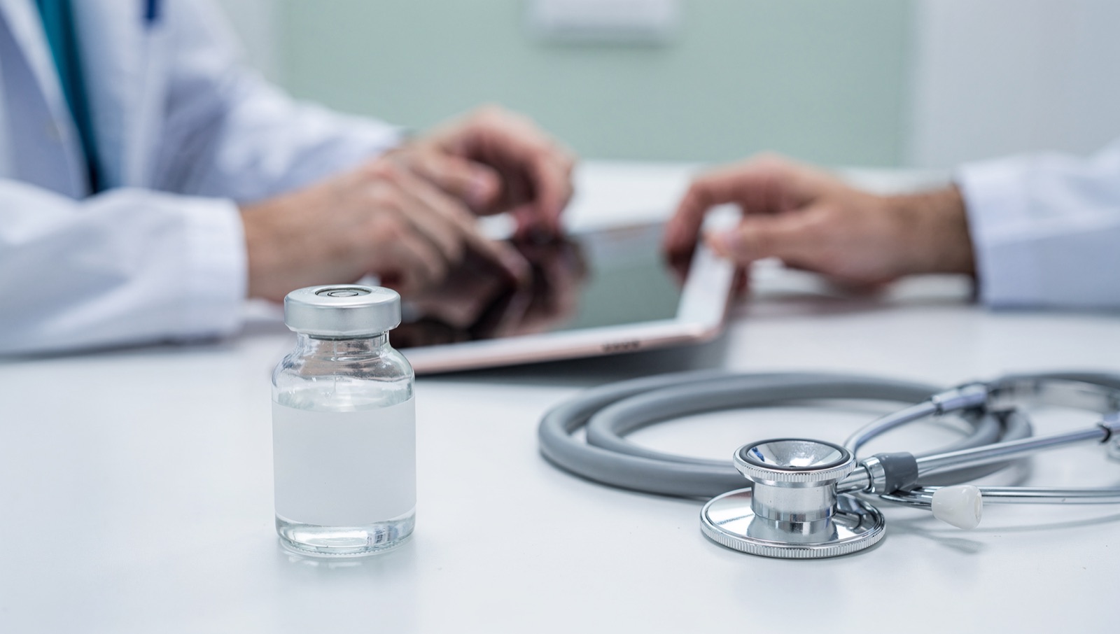 Medication vial and stethoscope on a clinic desk while a provider reviews research on a tablet