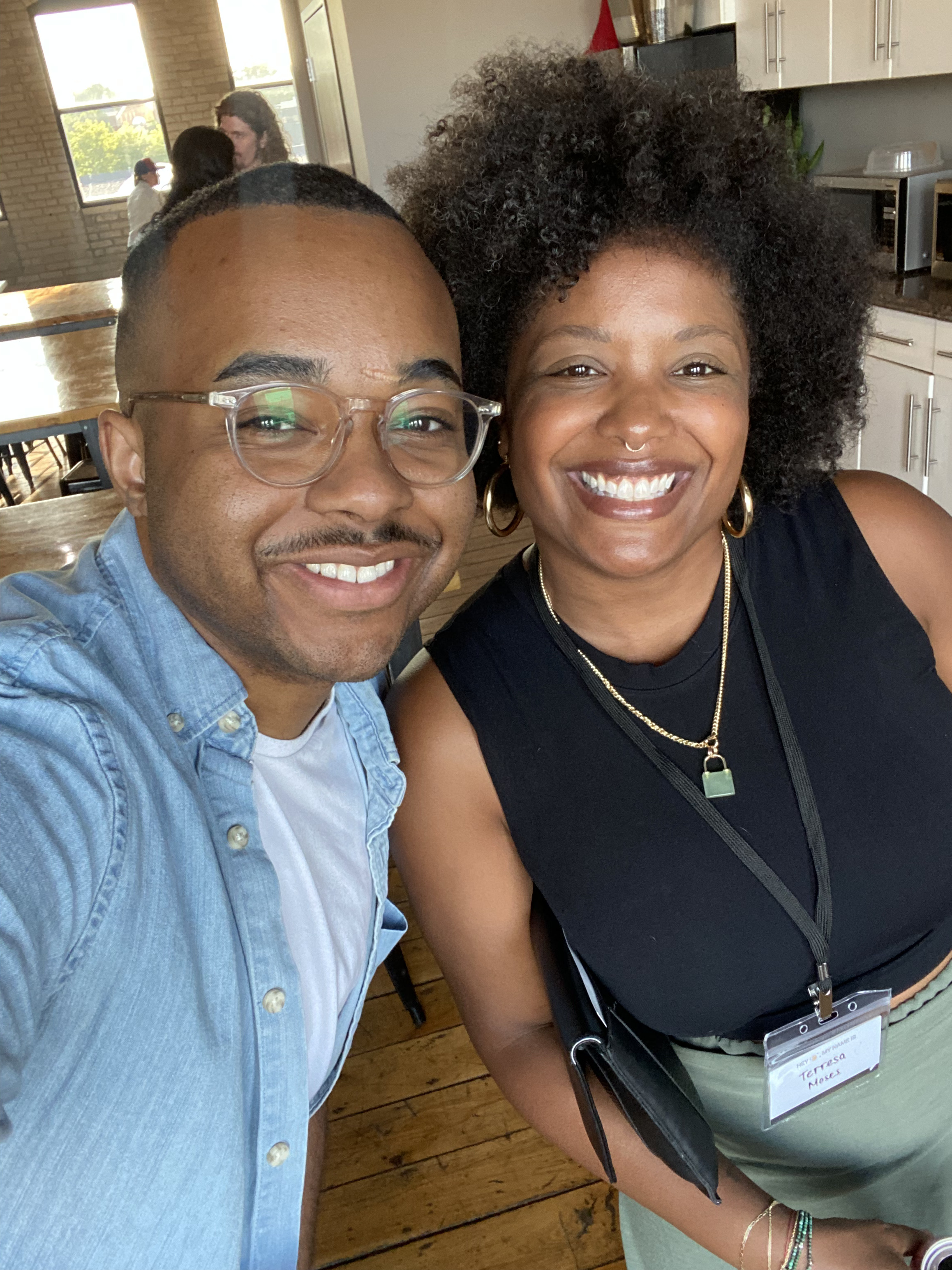 Smiling man in glasses and denim shirt taking a selfie with a woman wearing a black top, green pants, and a name tag reading Teresa Moses indoors.