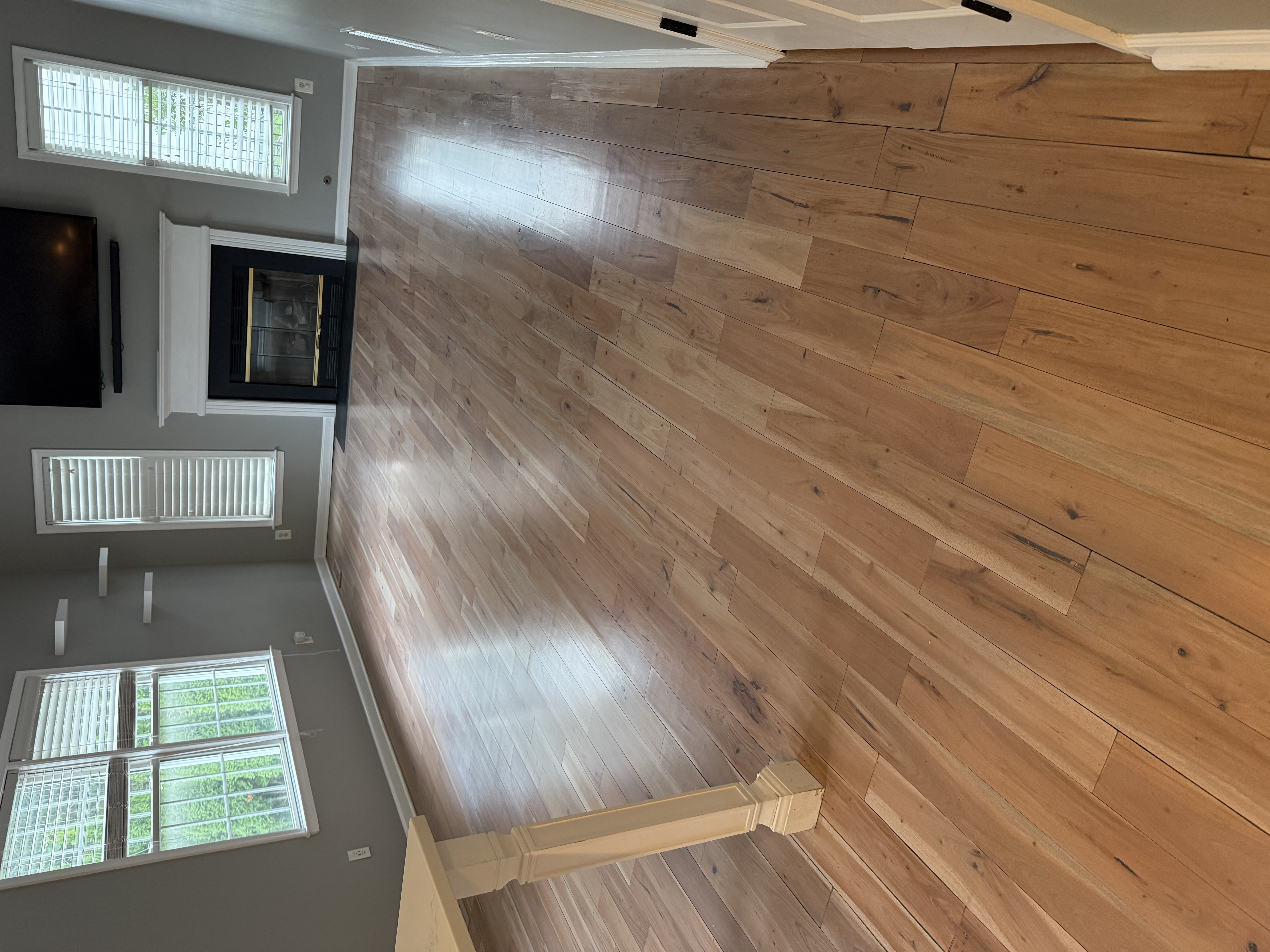 Empty room with light hardwood floors, gray walls, a white fireplace with a black TV mounted above, and windows with white blinds.