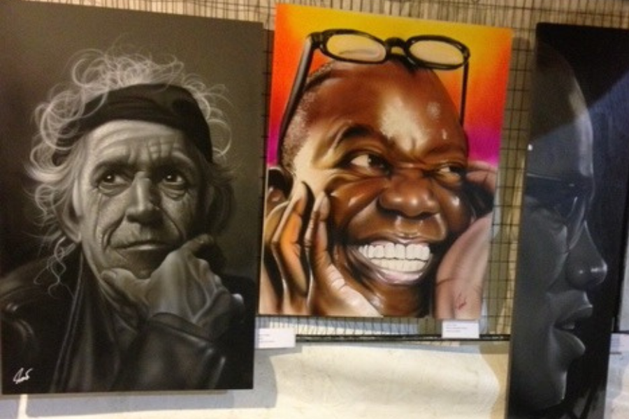 Three portraits displayed on a wall: a black-and-white painting of an older man with white hair and a contemplative expression on the left, a colorful painting of a smiling man with glasses on his head in the center, and a dark monochrome profile of a face on the right.