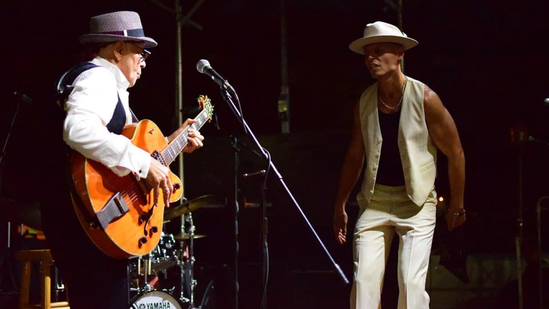 Musician playing an orange electric guitar on stage beside a man in a light vest and hat performing.