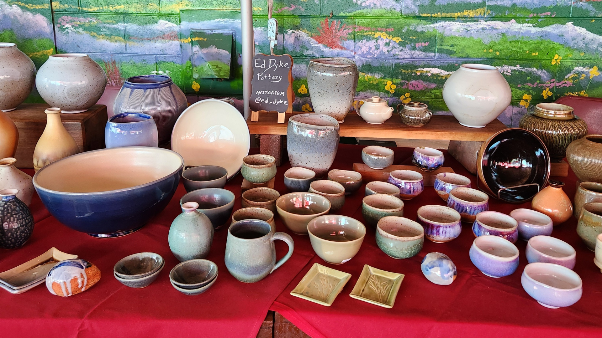 Table display of various handmade pottery pieces including bowls, mugs, vases, and small dishes on a red cloth with a colorful mural background and a sign reading 'Ed Dyke Pottery Instagram @ed_dyke'.