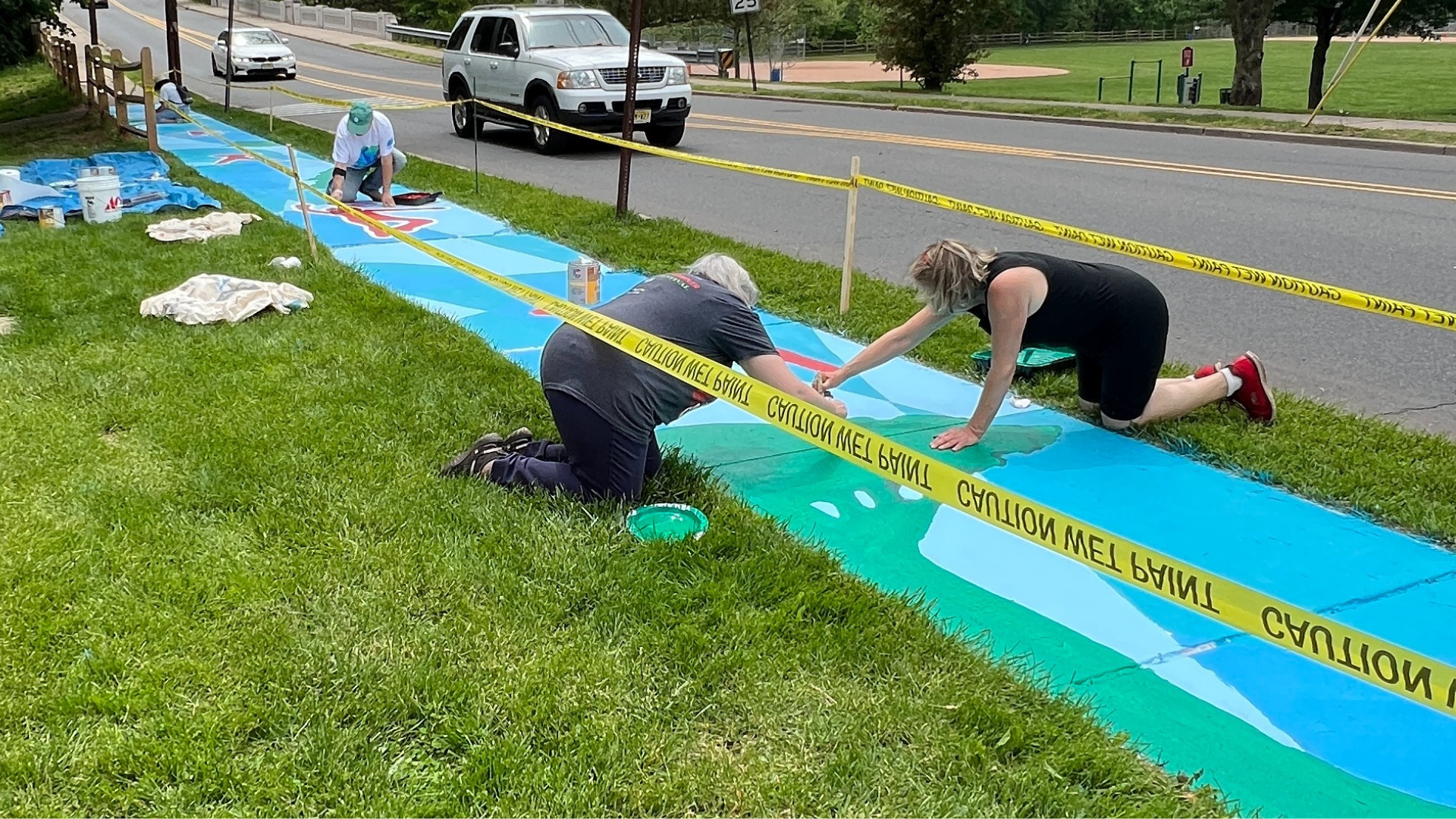 Three people kneeling on grass painting a long colorful mural on the sidewalk, surrounded by yellow caution tape.