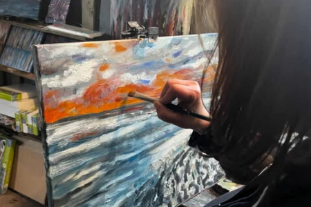 Person painting a landscape with a brush, featuring an orange sky and blue water on a canvas in an art studio.
