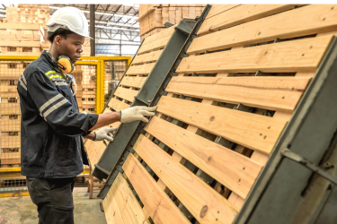 A worker wearing a hard hat and gloves inspects a wooden pallet secured upright in a warehouse fixture.