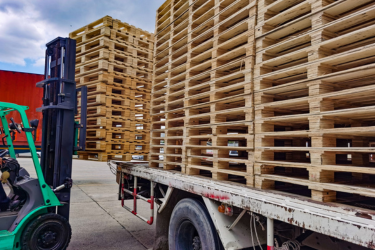 A forklift stands beside a flatbed trailer loaded with tall stacks of wooden pallets in an outdoor yard.