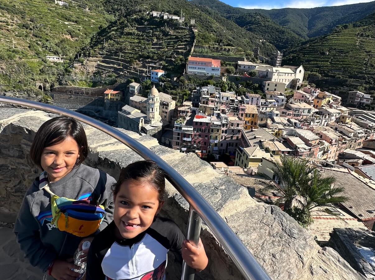 Two kids pose in front of a mountain village while slow traveling with family in Italy.