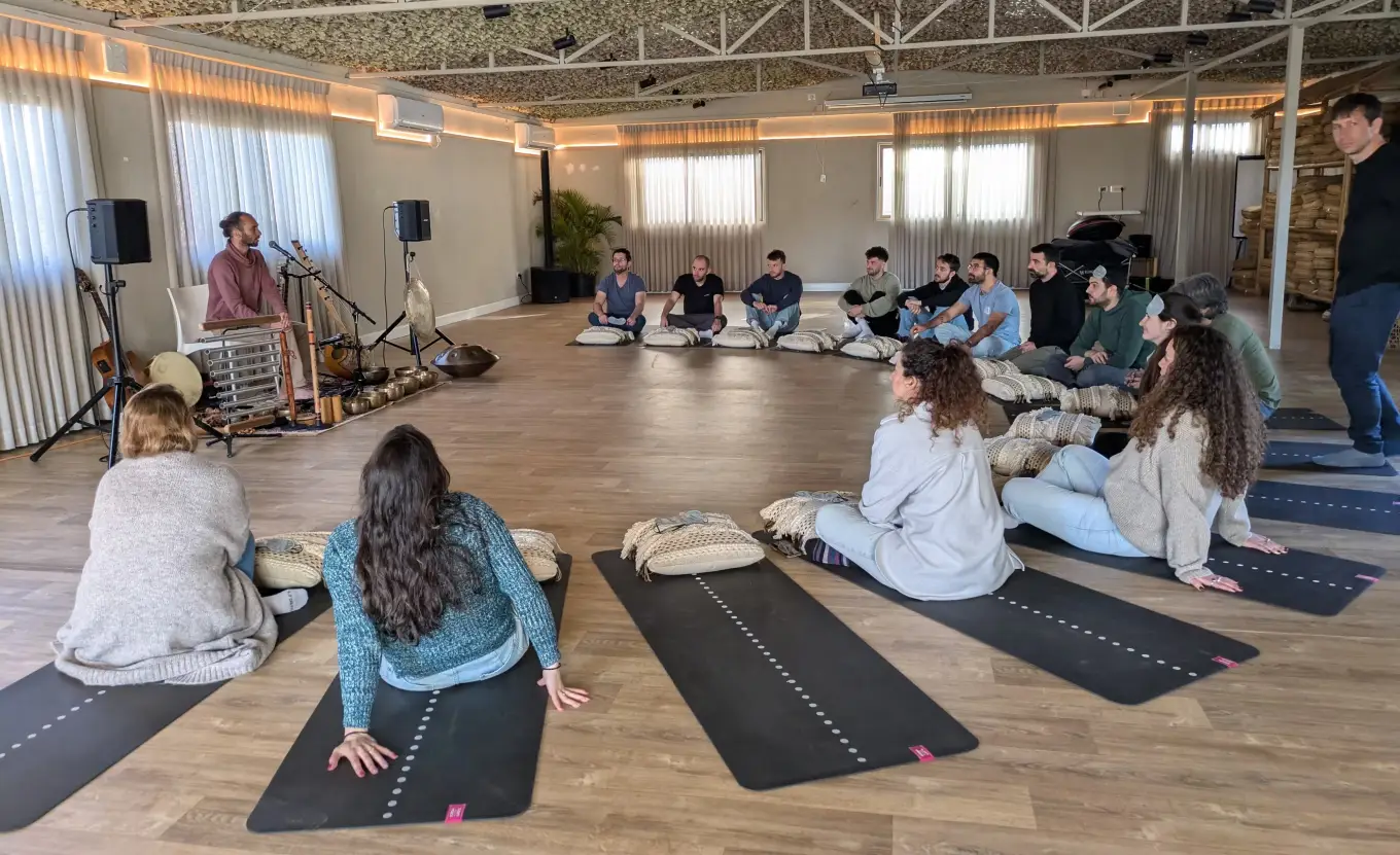 Group of people sitting in a spacious room on yoga mats and cushions arranged in a semicircle, listening to a man playing traditional musical instruments including a gong and various percussion instruments.