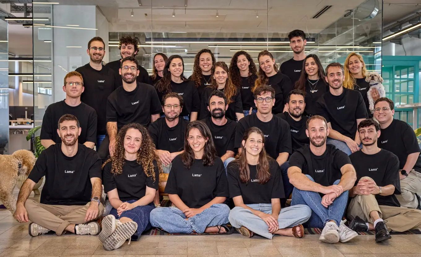 Group portrait of a diverse team of 26 people wearing matching black Lama t-shirts, seated and standing in an office with glass walls, smiling at the camera, with two small dogs present on the left and right sides.