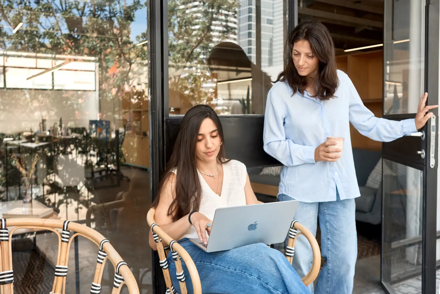 Two women at a cafe or office with glass doors; one woman wearing a white sleeveless top and jeans sits on a bamboo-style chair using a MacBook laptop while the other woman in a light blue shirt and jeans stands holding a cup and the door open.