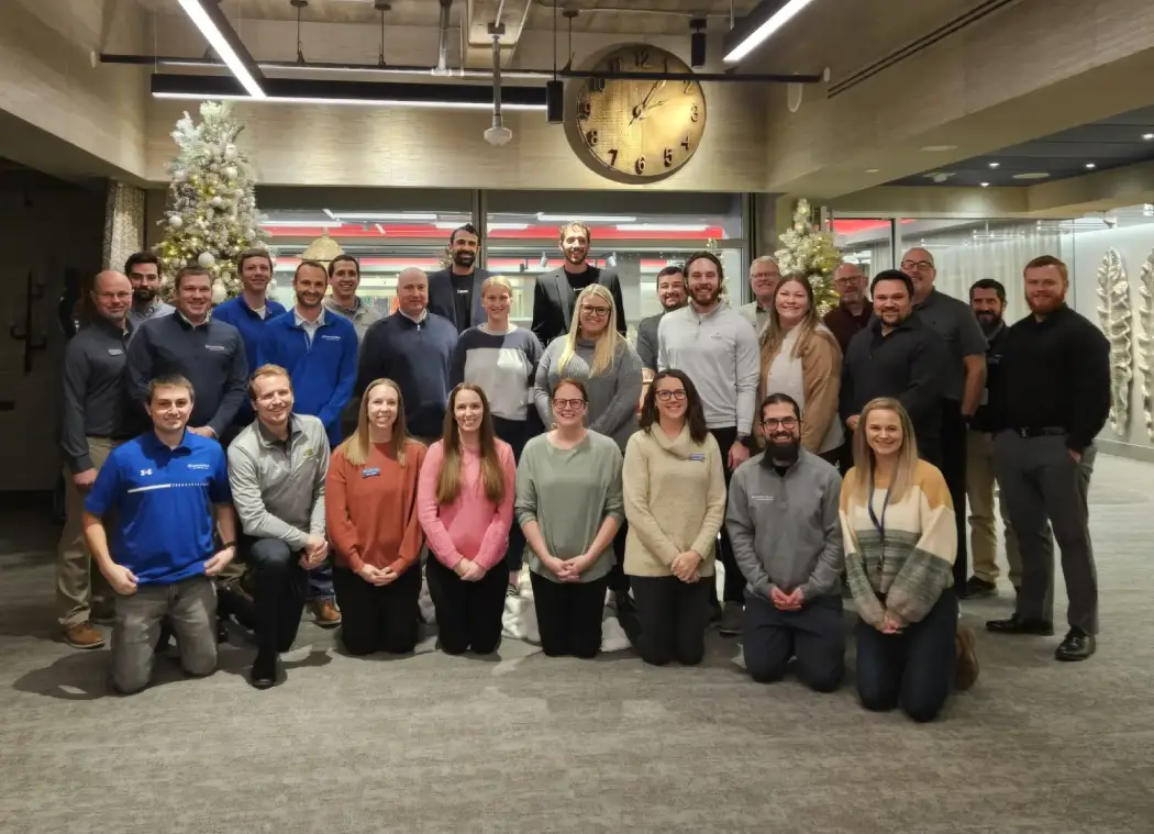 Group photo of 27 people posing indoors in three rows with two decorated white Christmas trees on either side and a large round clock showing 12:05 above them.