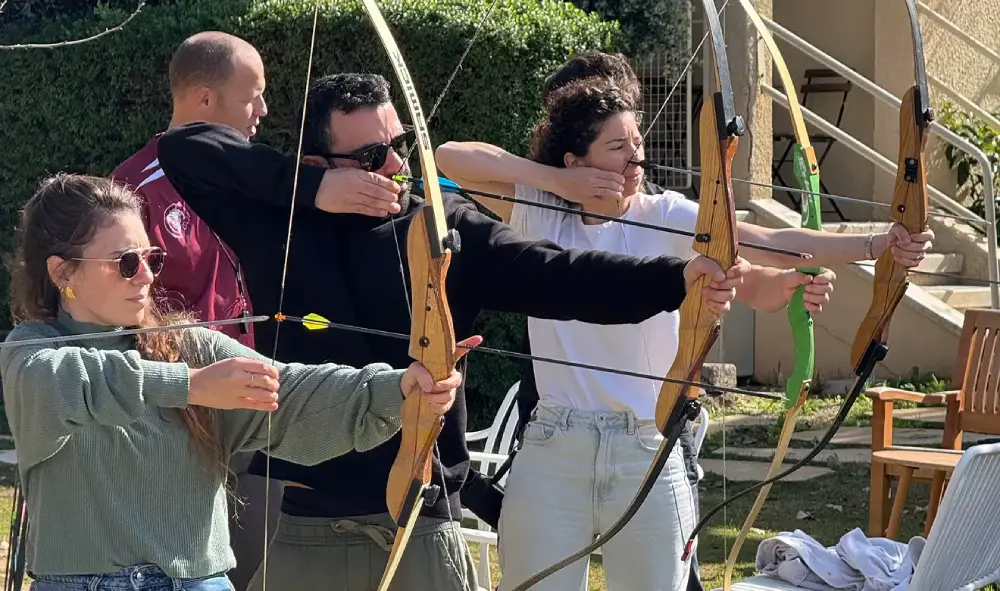 Three people outdoors aiming wooden recurve bows with arrows drawn, practicing archery on a sunny day.