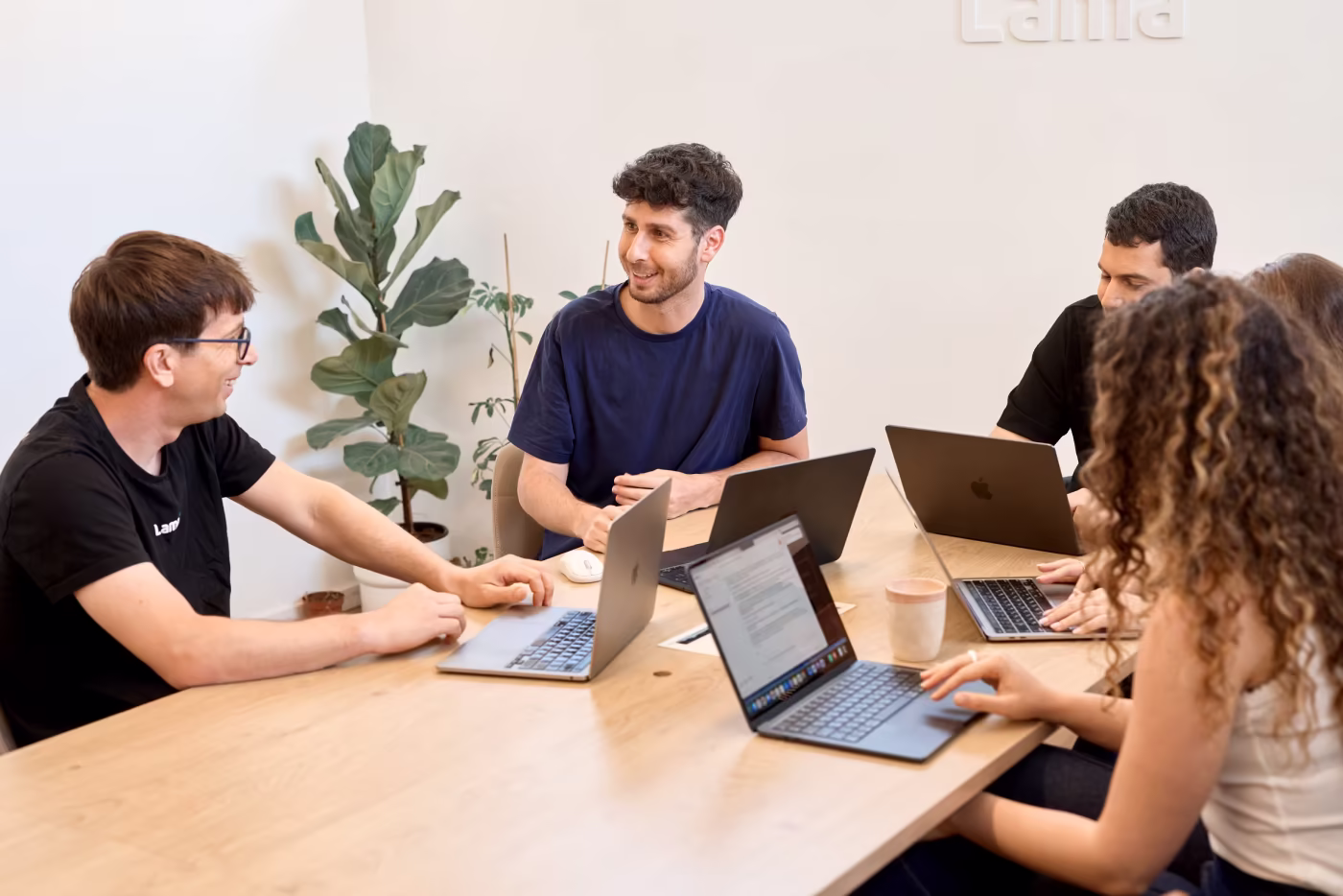 Four people sit around a wooden table in a meeting room with laptops open, engaged in conversation and collaboration, with a potted plant visible in the background.
