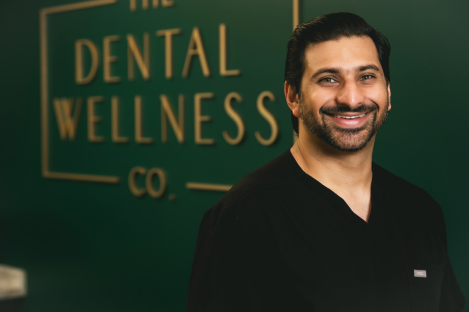 Smiling dentist in black shirt standing in front of The Dental Wellness Co. green branded wall.
