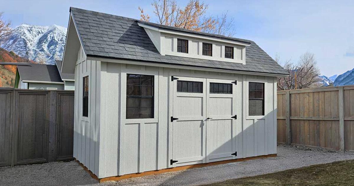 White custom storage shed with dormer roof, double doors, windows, and mountain backdrop
