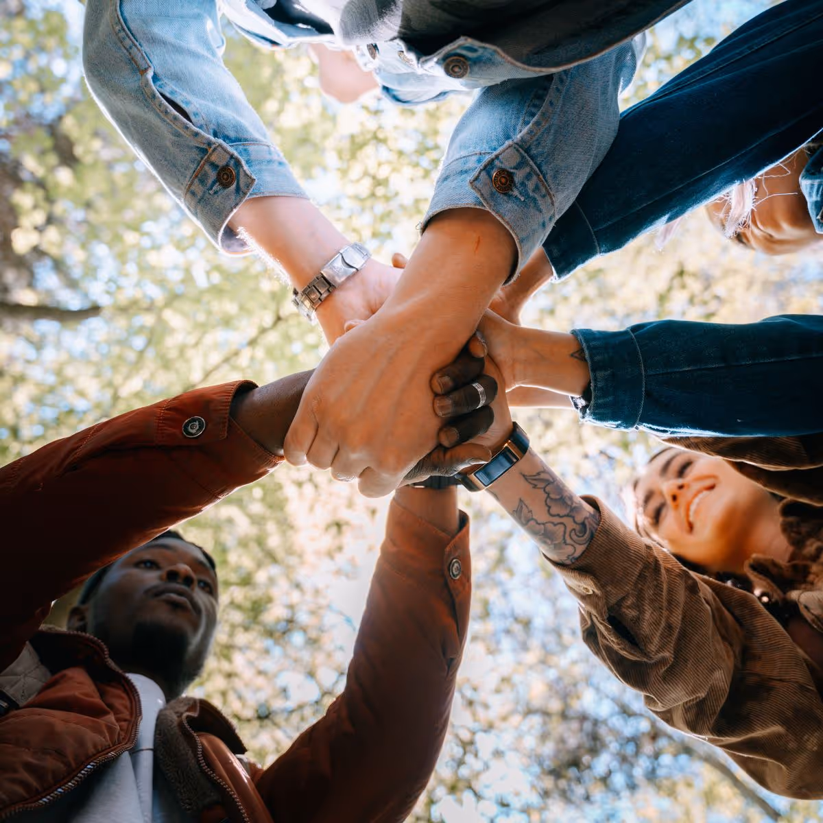 A group of co-workers with their hands connected signifying unity. This shot is from the ground looking up at their hands and faces, trees and sky are above.