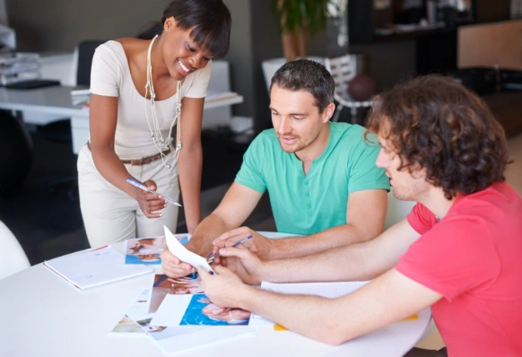 Three people collaborating around a table, reviewing documents and photos in an office setting.