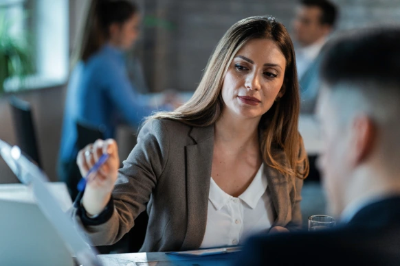 Businesswoman in a blazer attentively discussing work with a colleague in an office setting.