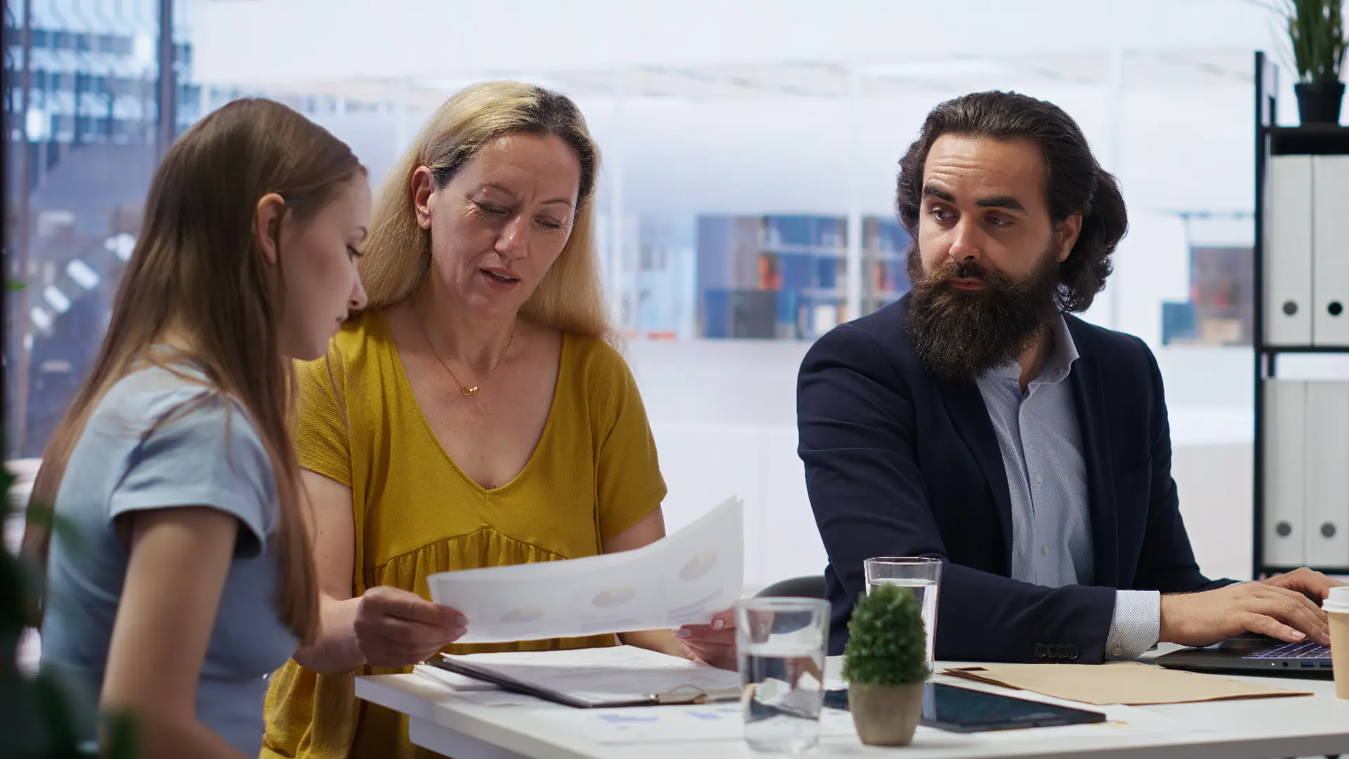 Three professionals at a desk reviewing documents and using a laptop in a modern office setting.