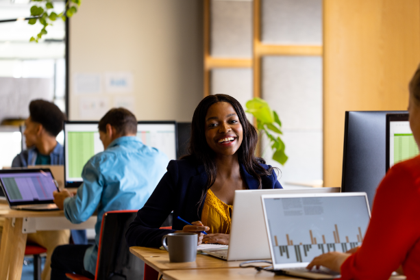 Smiling woman in a mustard top and dark blazer sitting at a desk with a laptop, interacting with a colleague in an office setting.