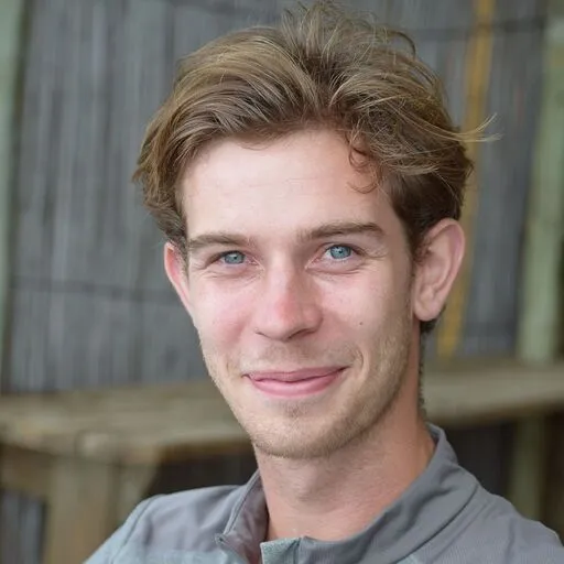 Young man with light brown hair and blue eyes smiling slightly in an outdoor setting.