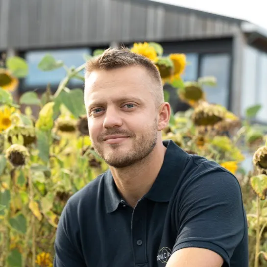 Smiling man with short hair and a beard wearing a black polo shirt, sitting in front of a field of sunflowers.