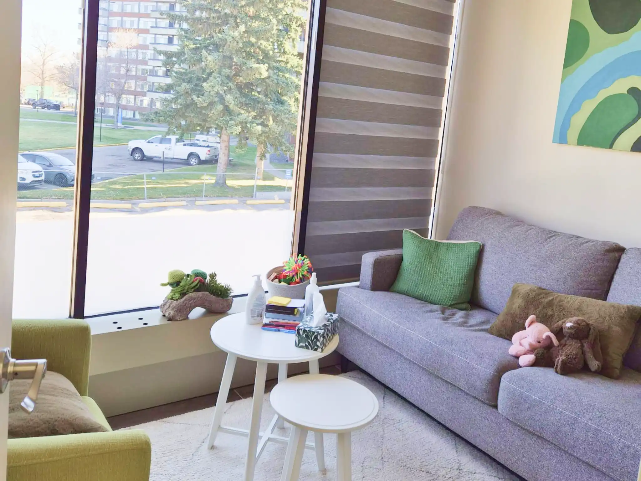 Bright therapy room with large window, gray couch with green pillows and stuffed animals, white tables with tissues and supplies, and a green armchair.