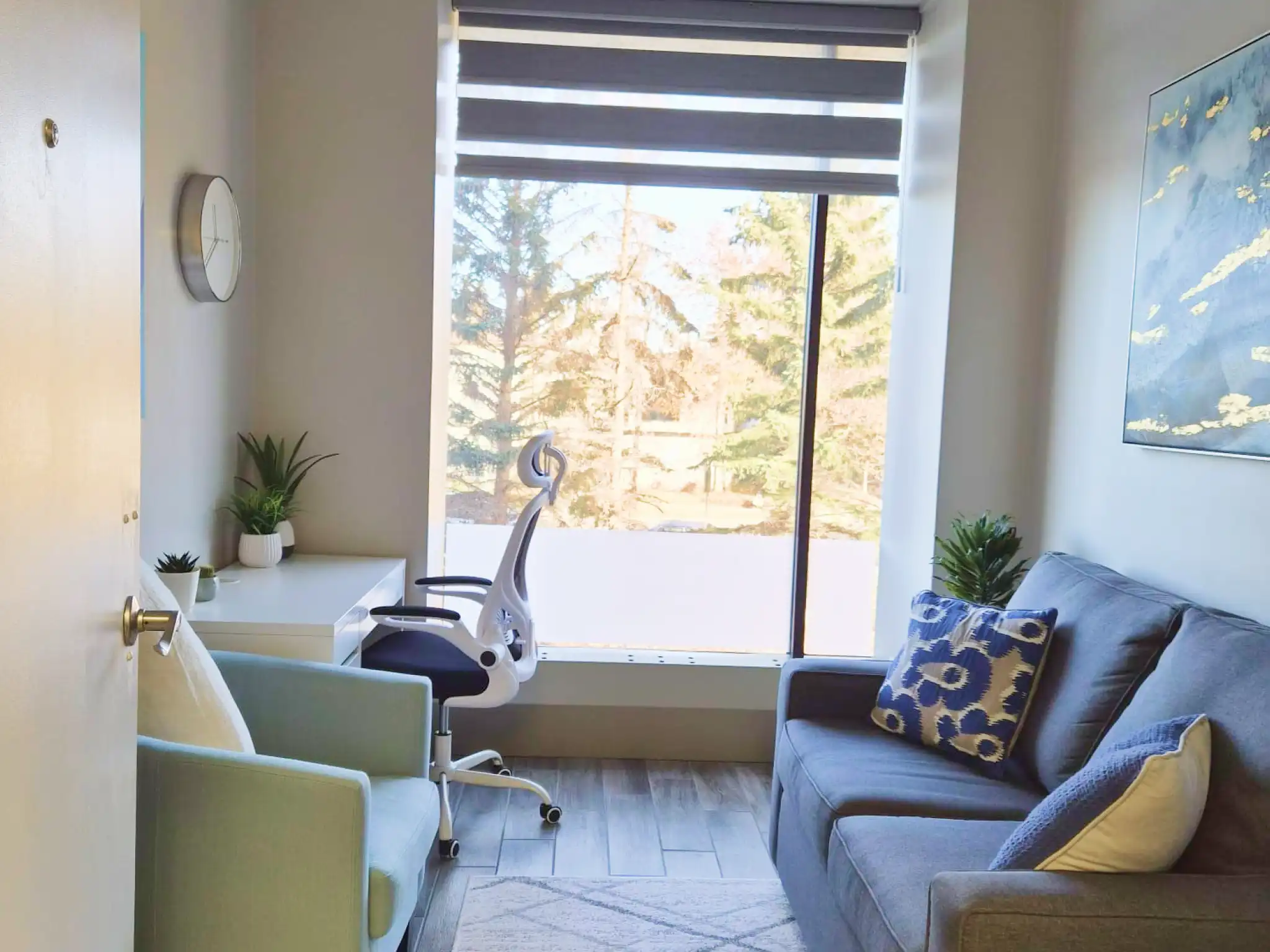 Bright therapy room with a large window, gray sofa with patterned pillows, light blue armchair, white desk chair, plants, and a beige geometric rug.