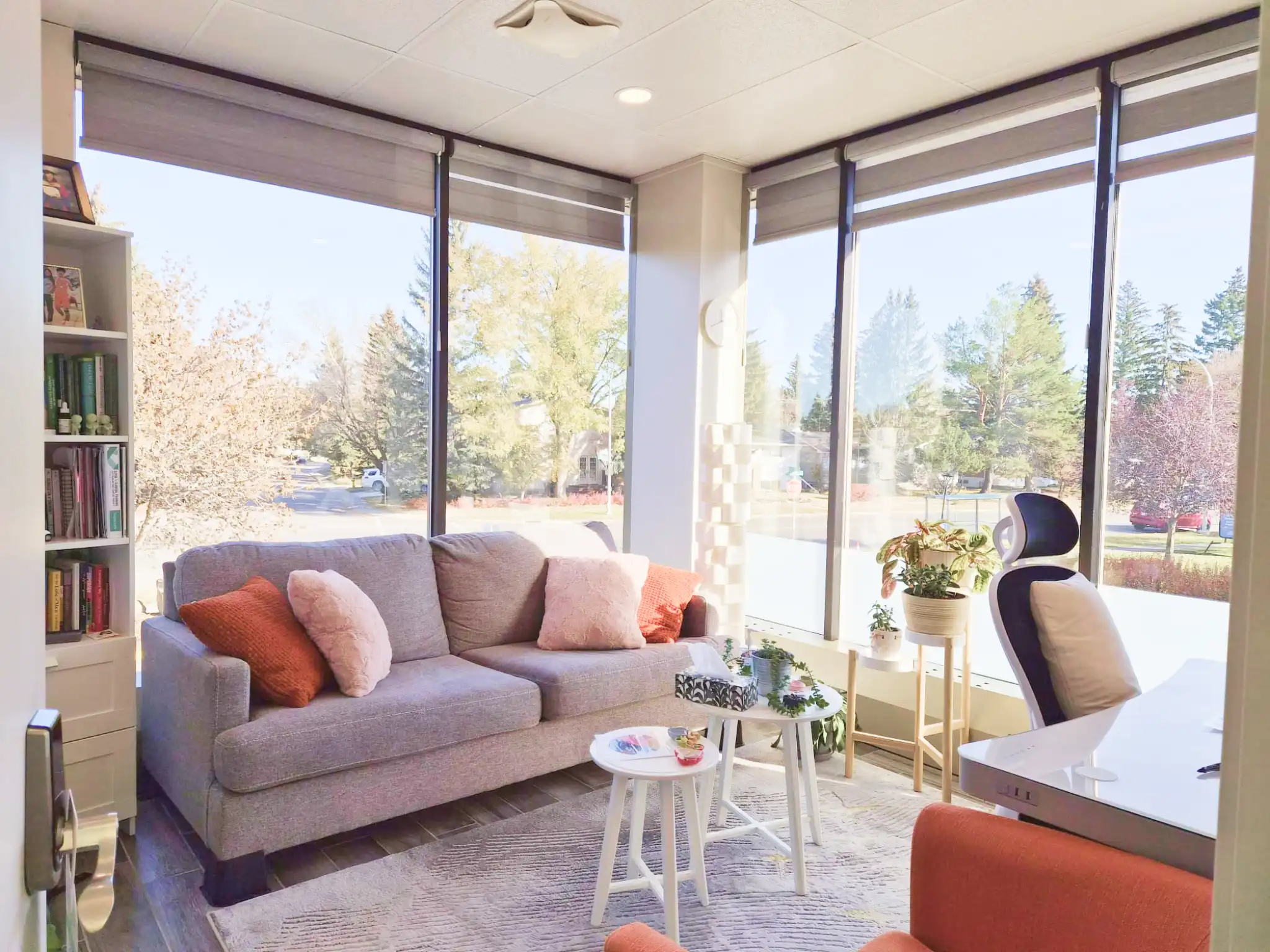 Bright therapy room with large windows, a gray couch with orange and pink cushions, white nesting tables, and a black ergonomic chair next to indoor plants.