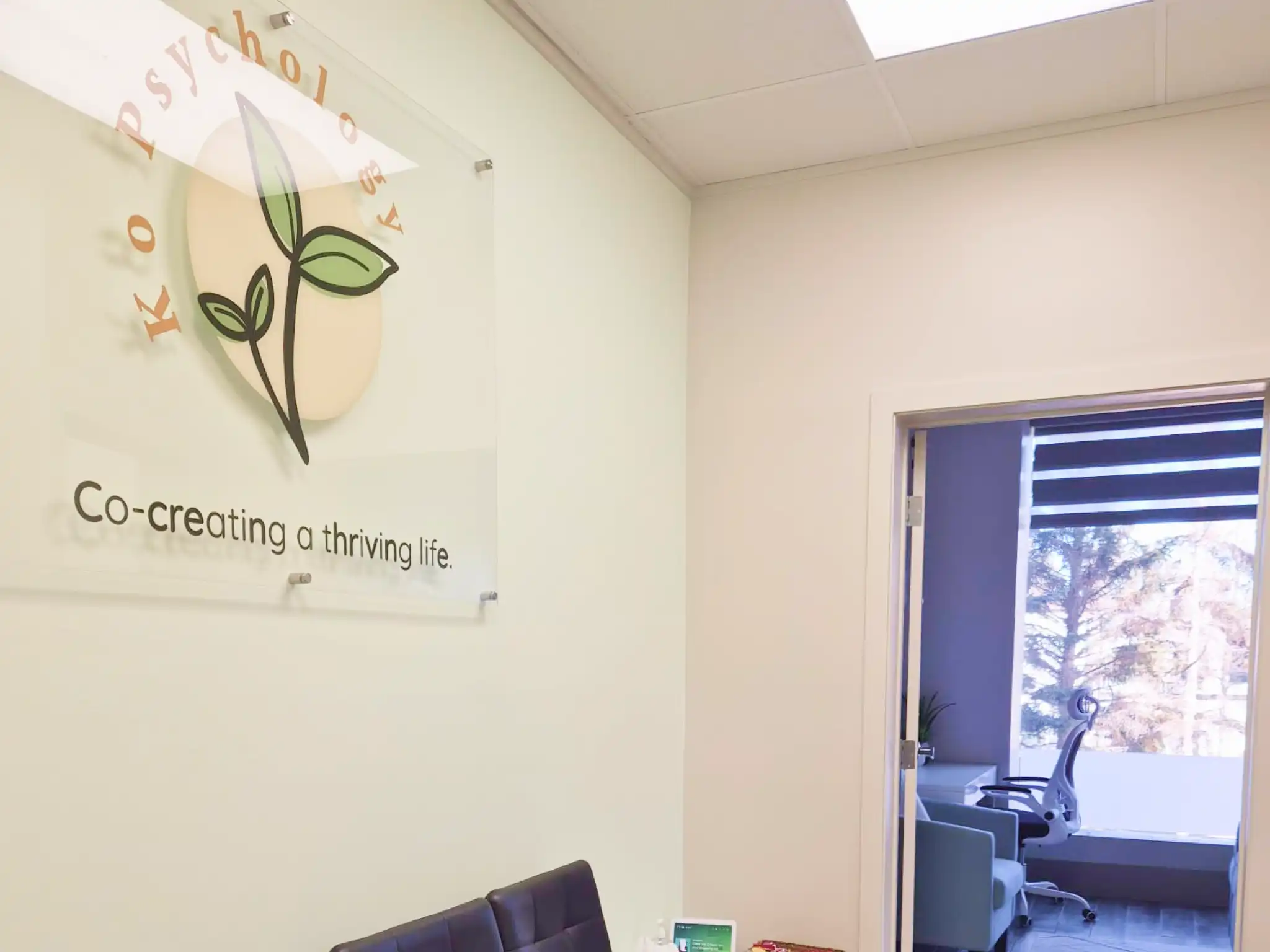 Waiting area with three black cushioned chairs, a small white table with tissues and a basket, and a doorway leading to an office with a chair and window.