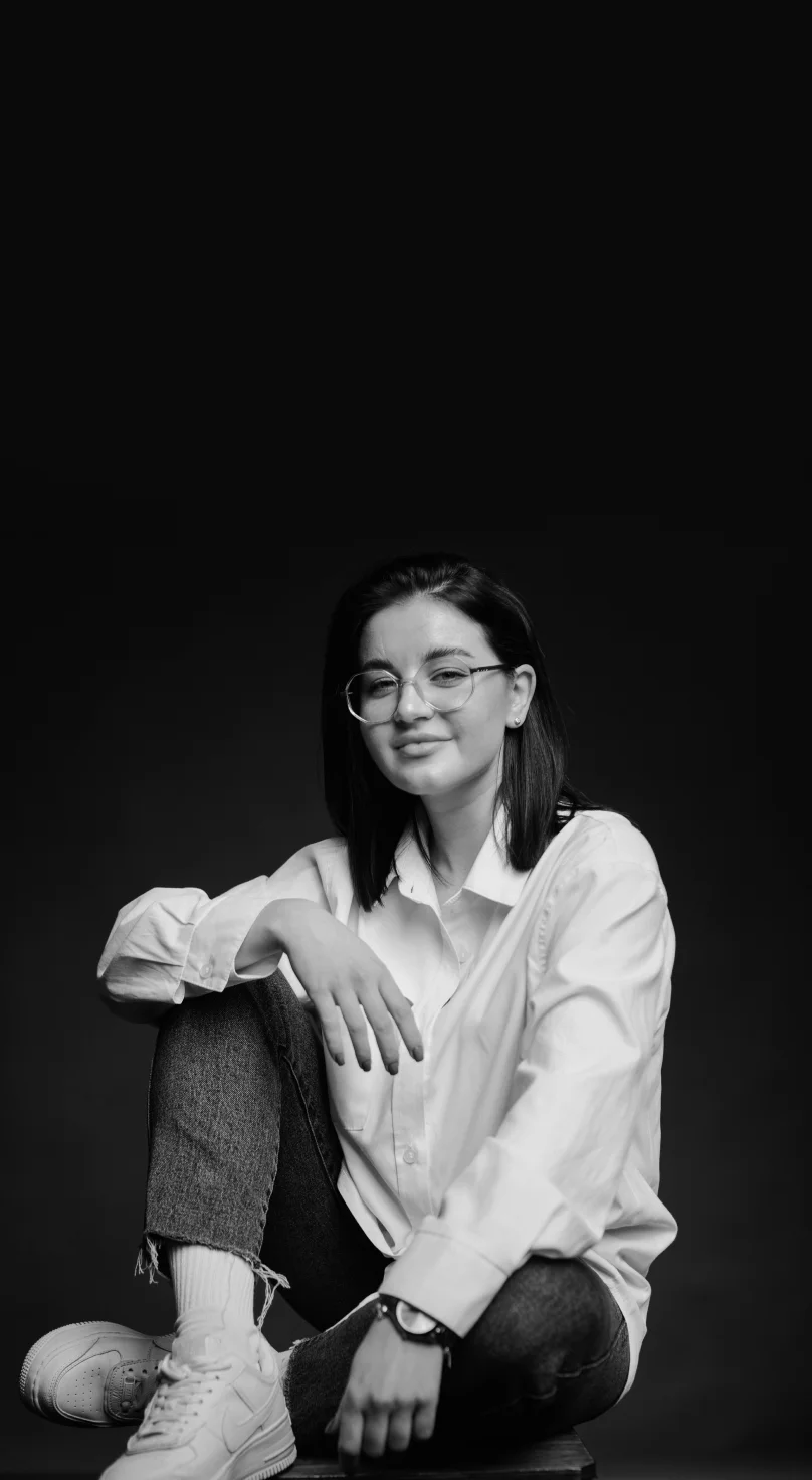 Young woman with glasses sitting cross-legged wearing a white shirt, jeans, and sneakers against a dark background.