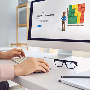 Mitarbeiter absolviert interaktives Risikomanagement-E-Learning am Desktop-Monitor. Die Lektion zeigt Schritt 2 – Bewertung mit einer modernen 3D-Risikomatrix (Schadensausmaß und Eintrittswahrscheinlichkeit). Nahaufnahme von Händen auf der Tastatur am Schreibtisch mit Brille.