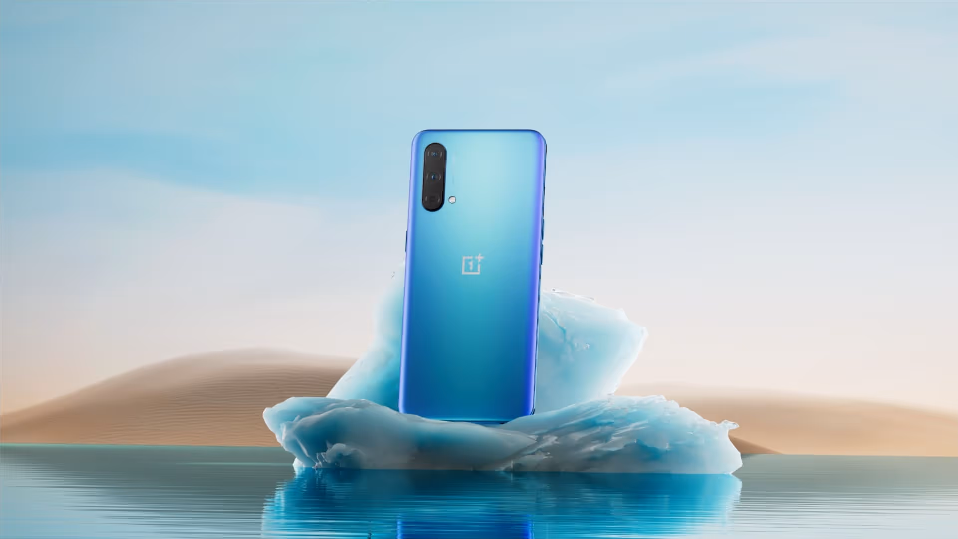 OnePlus device sits on an ice block in water against a sandy dune background.
