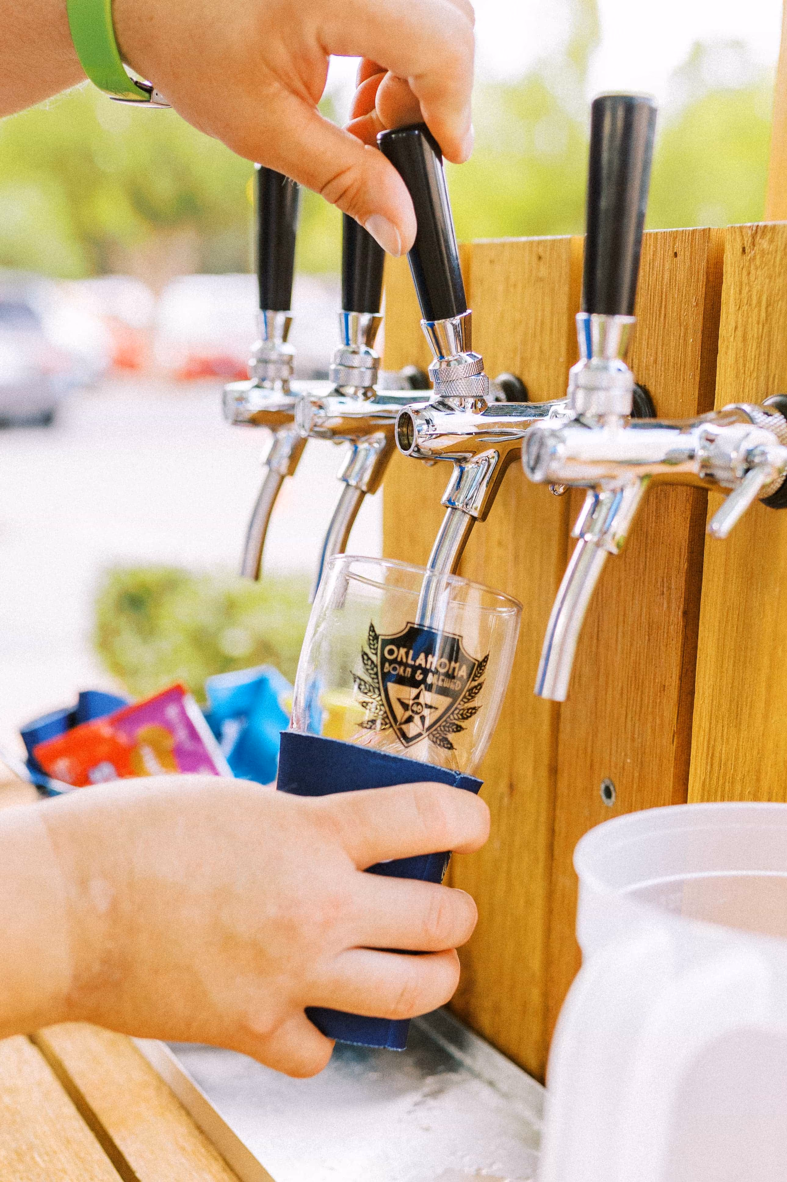 A close-up photo of a brewer pouring beer from a four-tap system into an Oklahoma Born & Brewed branded sample glass.