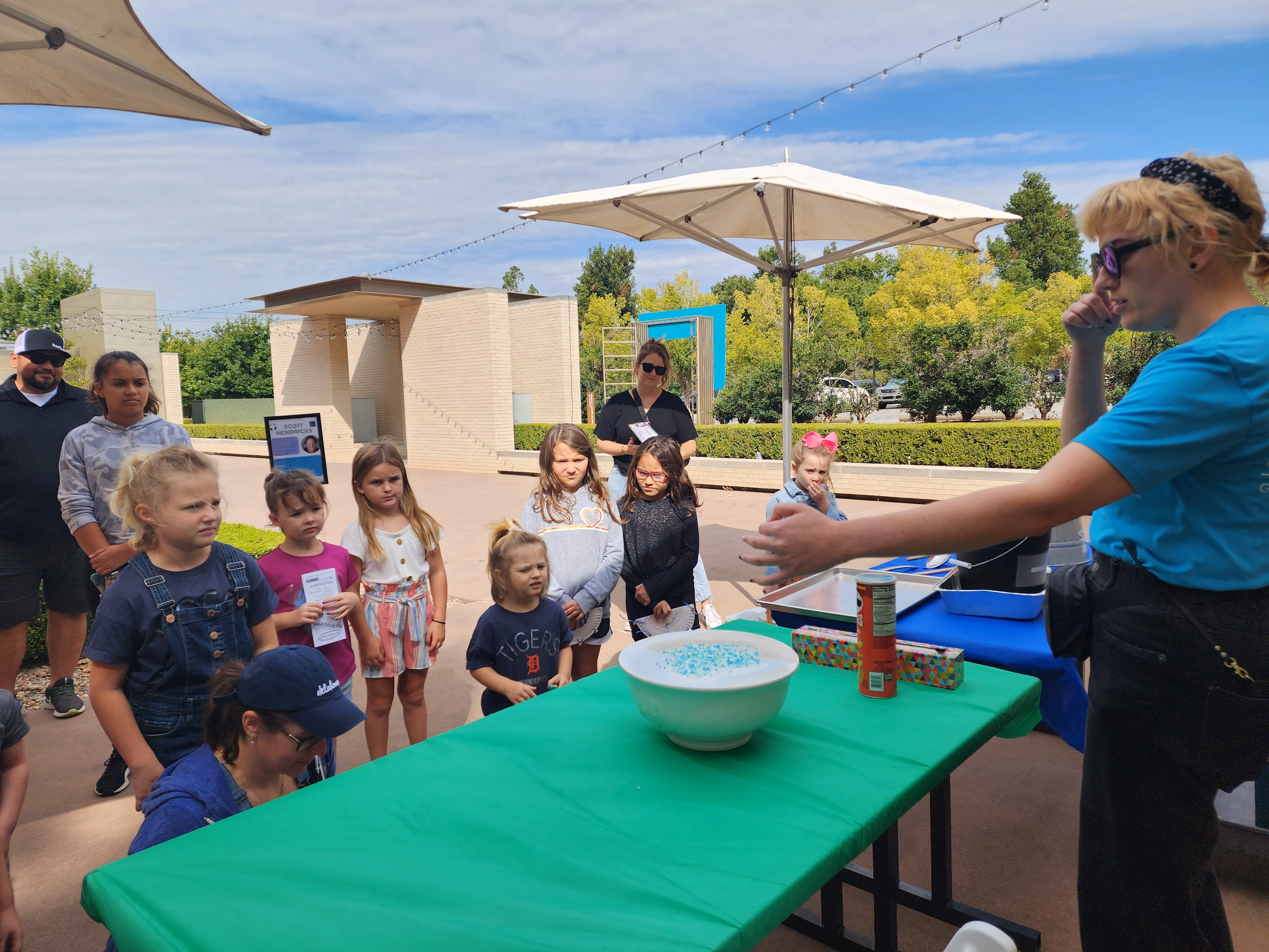A Museum educator is demonstrating that sound is vibration using a bowl with plastic wrap over it and blue-dyed grains of rice on top. A group of young student watch the demonstration in the Museum courtyard.