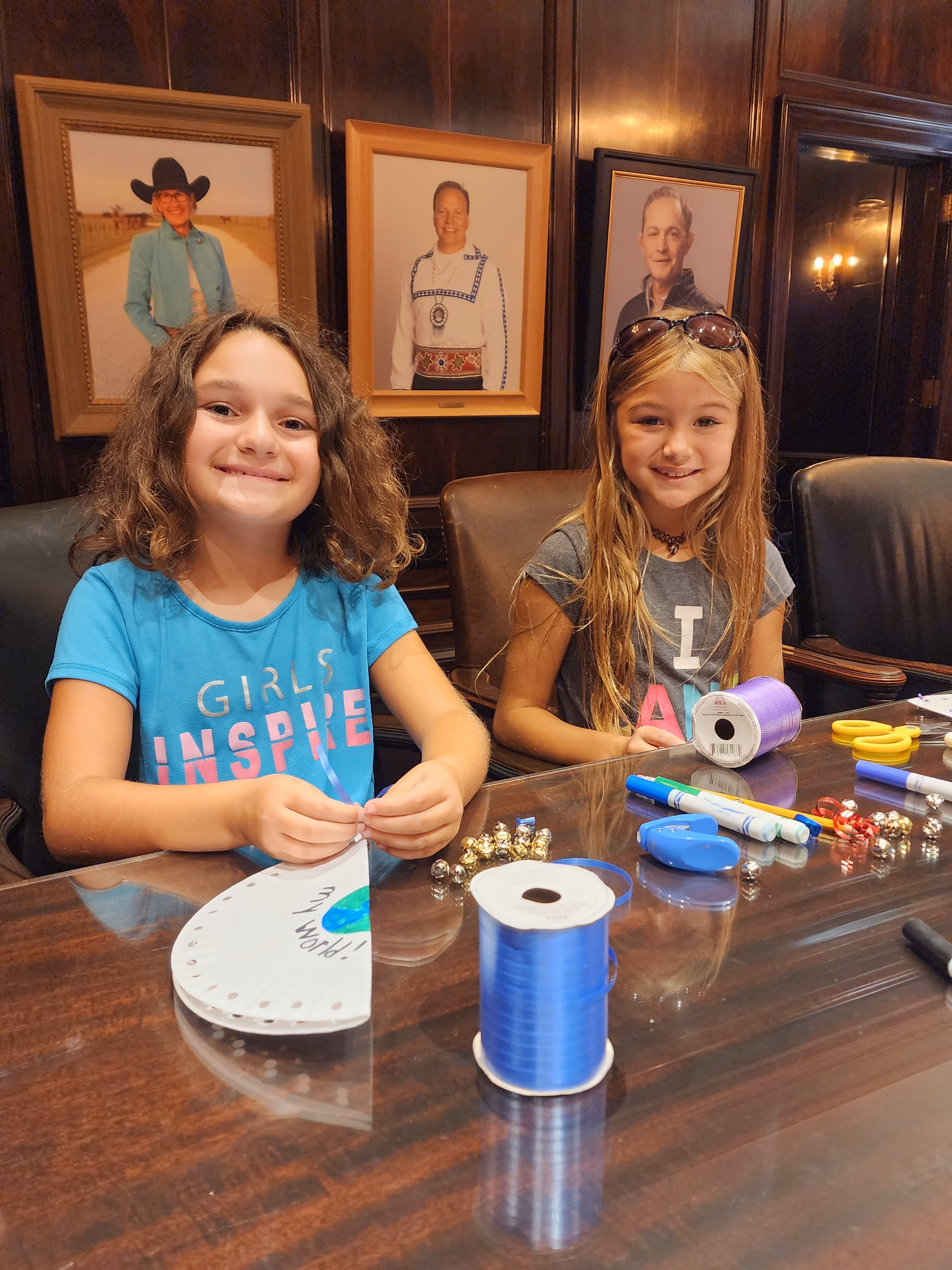 Two female students sit at a table while making paper plate tambourines out of ribbon and jingle bells.