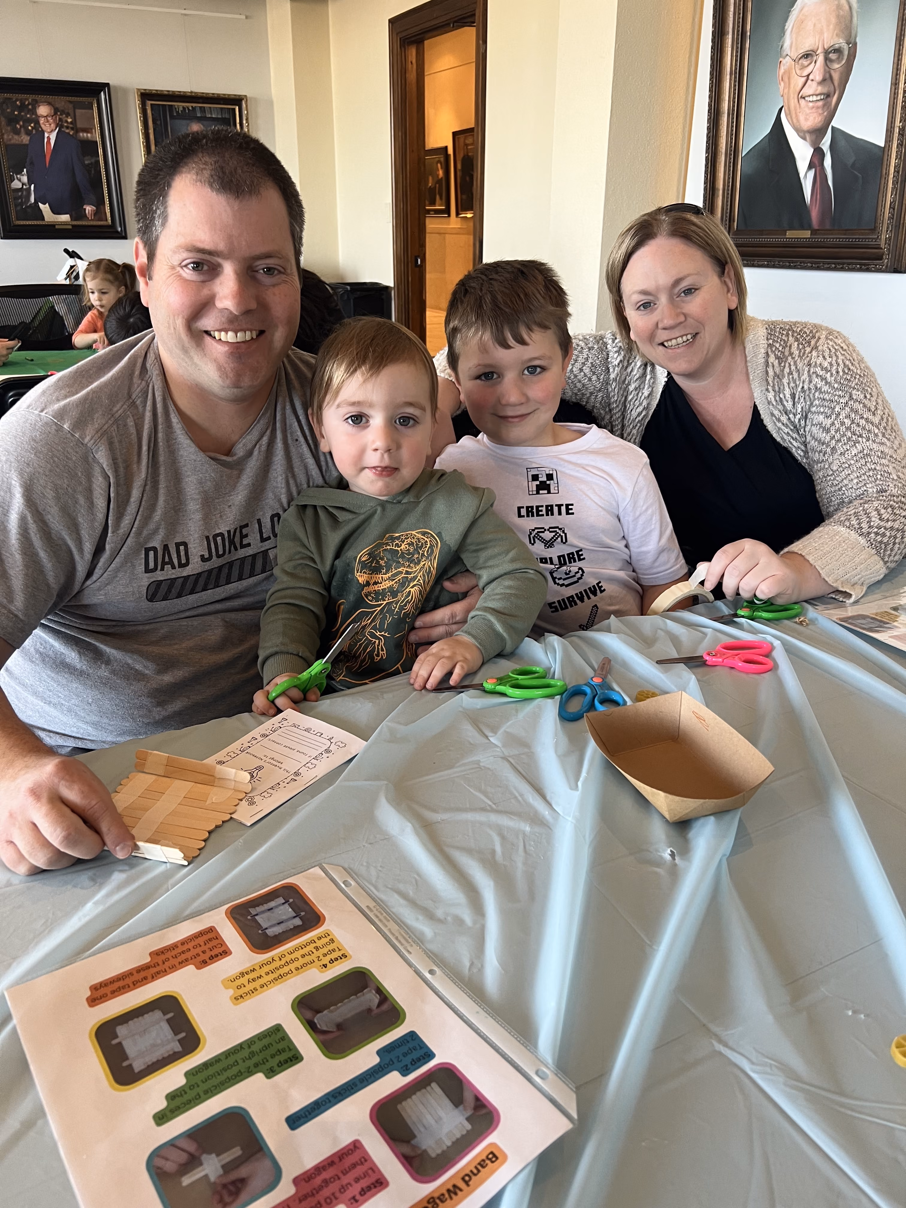 A family of four, two parents and two young children, sit at a craft table while making the rubber band-powered covered wagons out of popsicles and rubber bands.