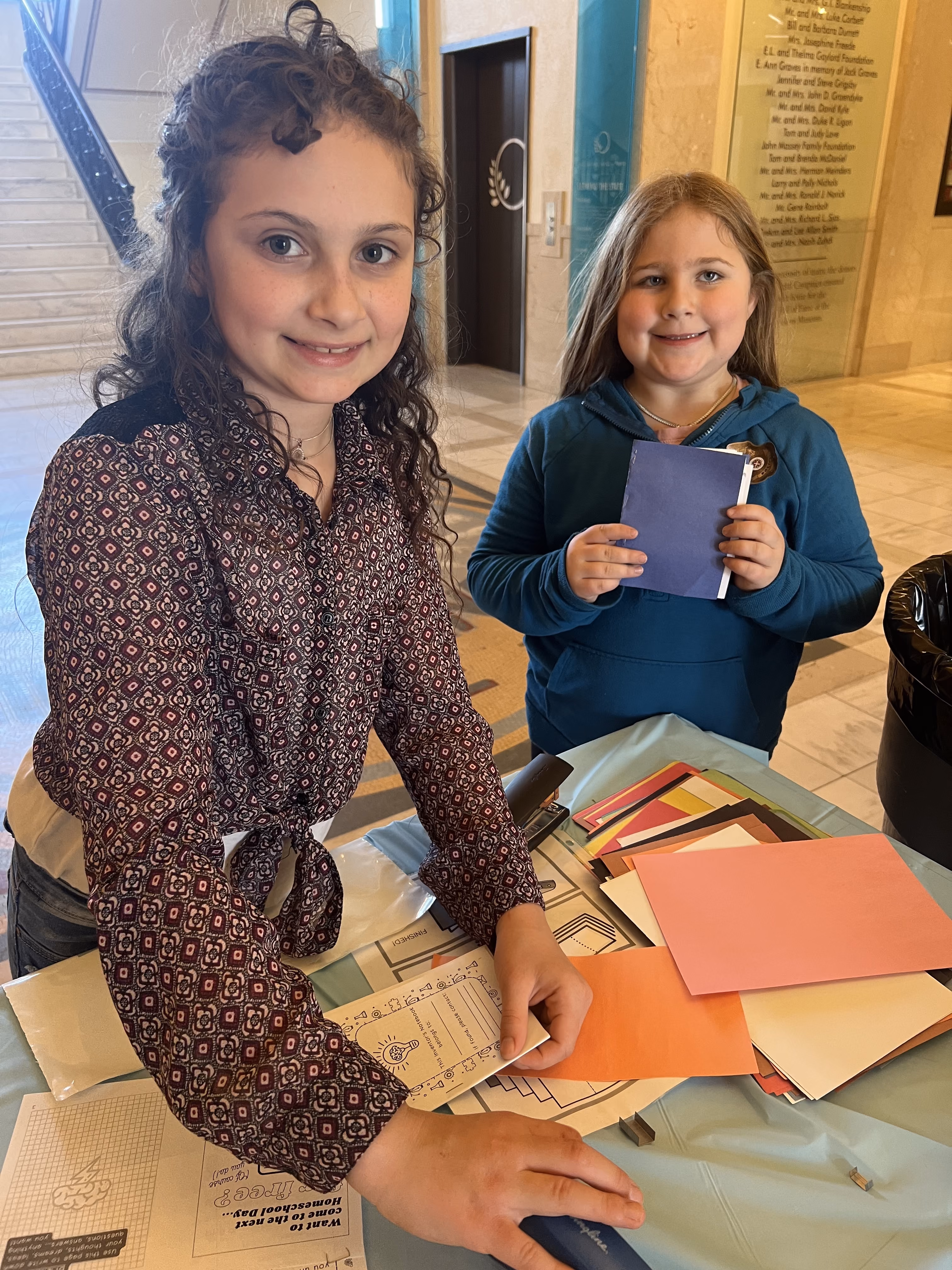 Two female students use construction paper to create Homeschool Day passports, which can be stamped as they complete that day's activities.
