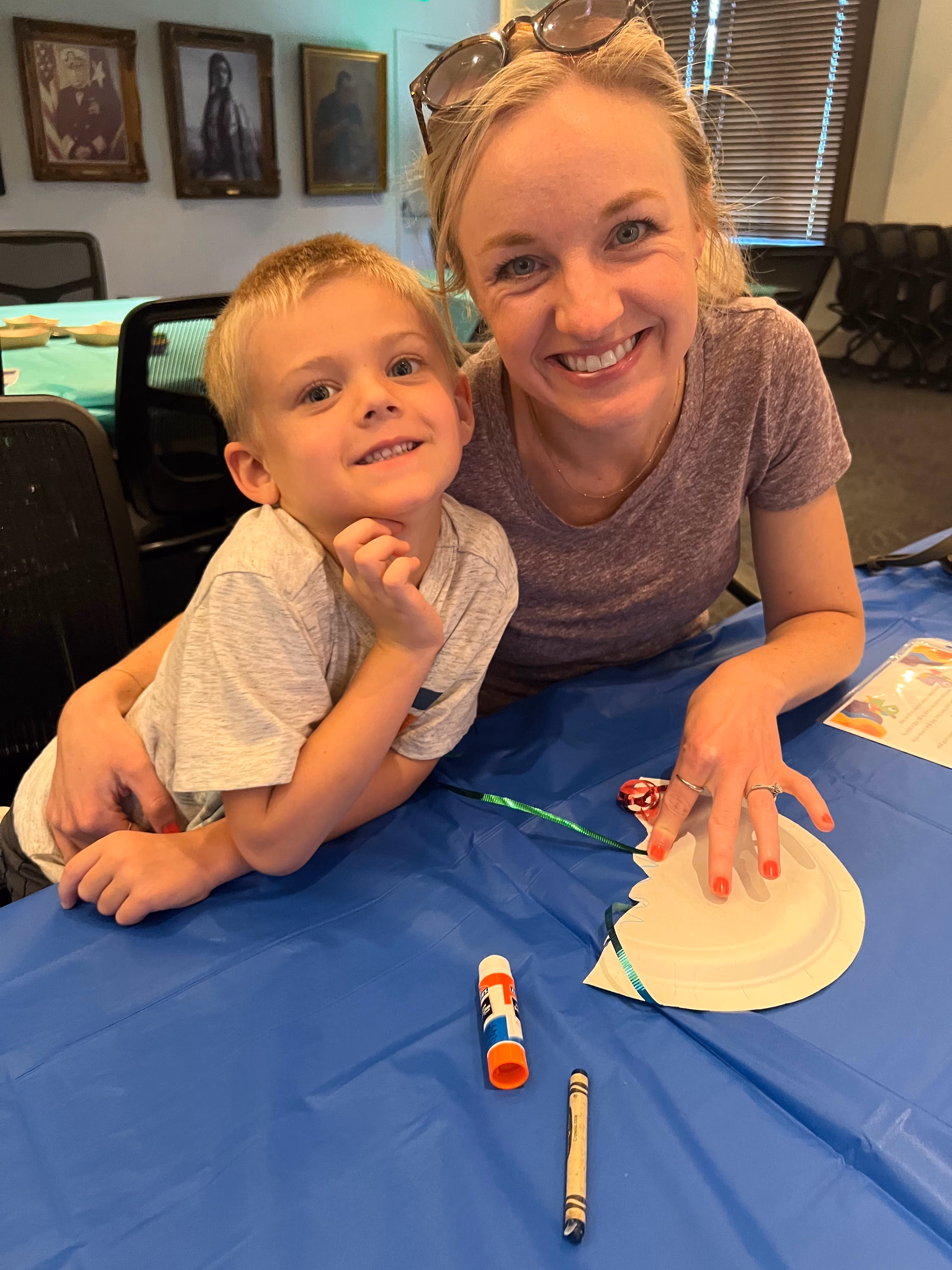 A mother and young son sit at a table, making a craft out of a paper plate.