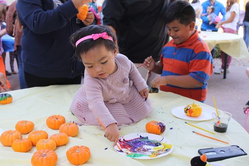 A toddler girl sits on a round craft table, surrounded by mini pumpkins. She uses a small brush to paint on a paper plate in front of her.