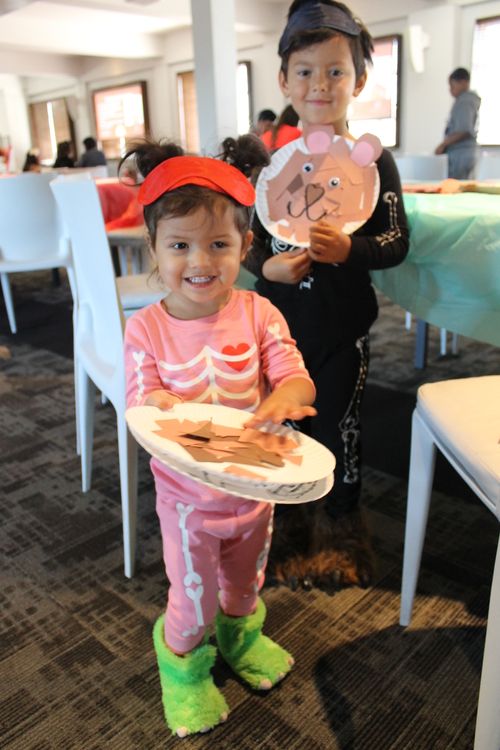 Two children dressed in skeleton pajamas, show the teddy bear face craft they made out of paper plates and torn up pieces of paper.