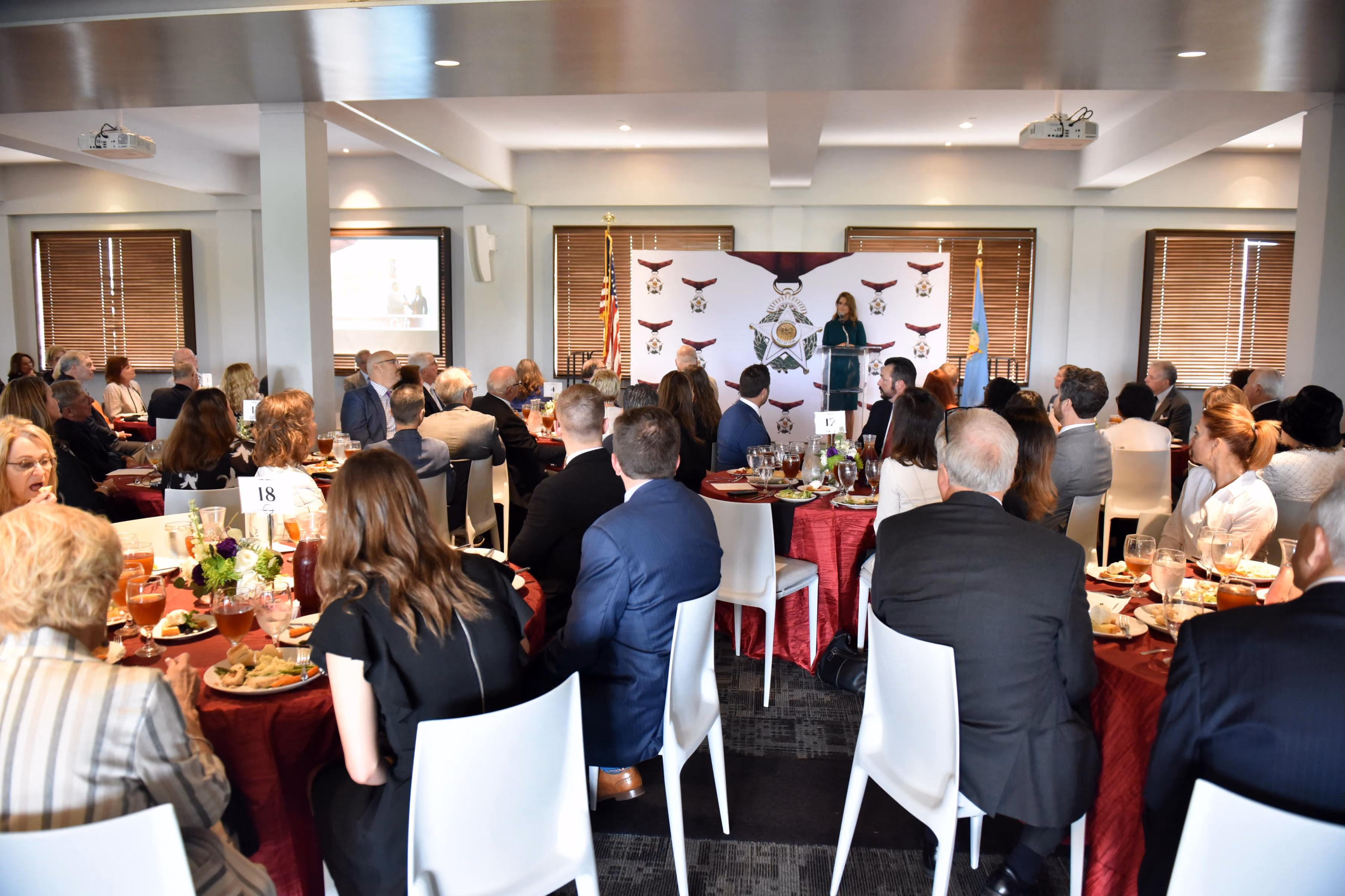 Guests seated at tables in a large event hall enjoy a meal while a woman at a podium at the front of the room gives a presentation, which is being projected onto a screen a few yards away. 
