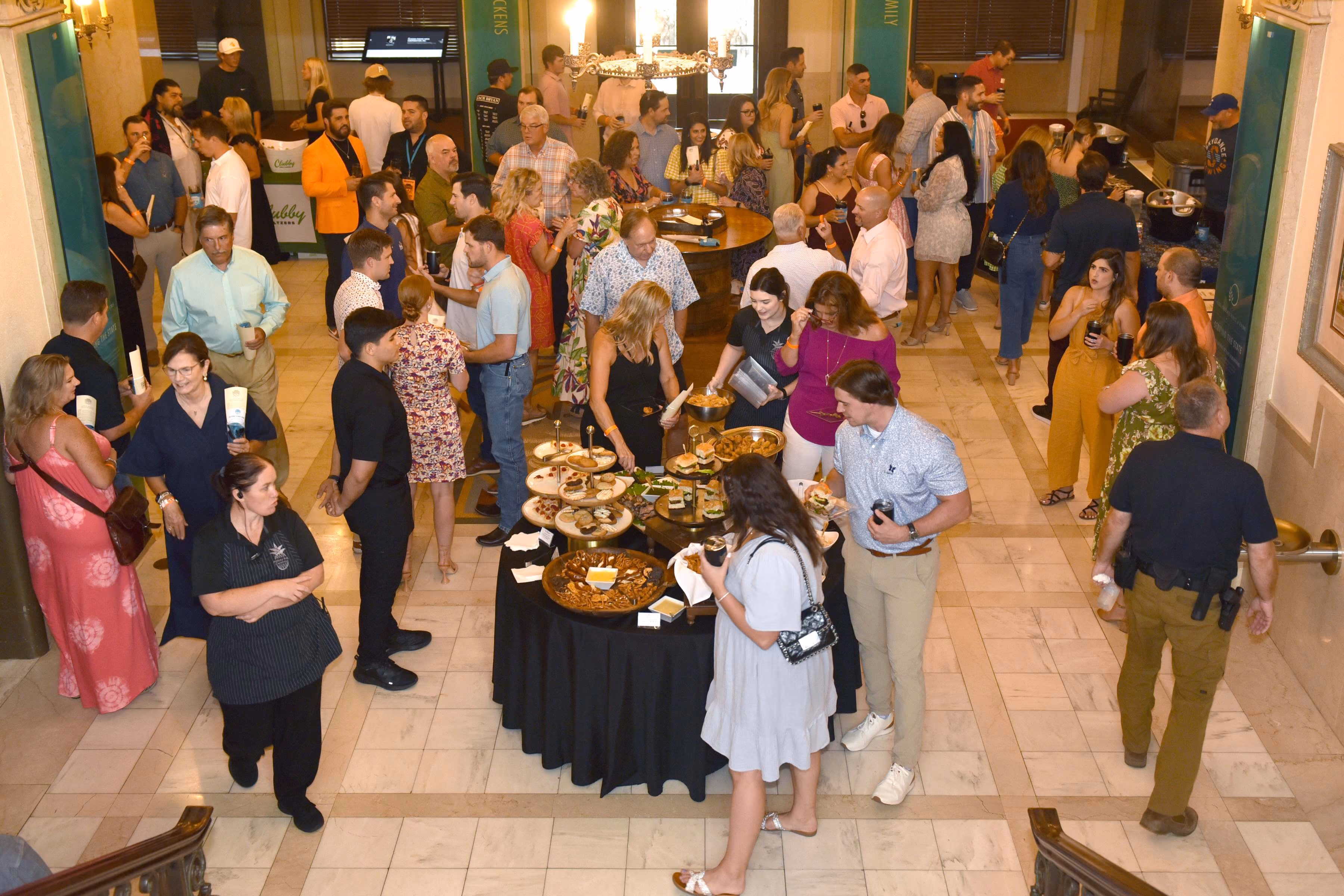 Guests gather and mingle in a marble-tiled foyer, enjoying drinks from several visible drink booths and filling plates at a table of appetizers. 