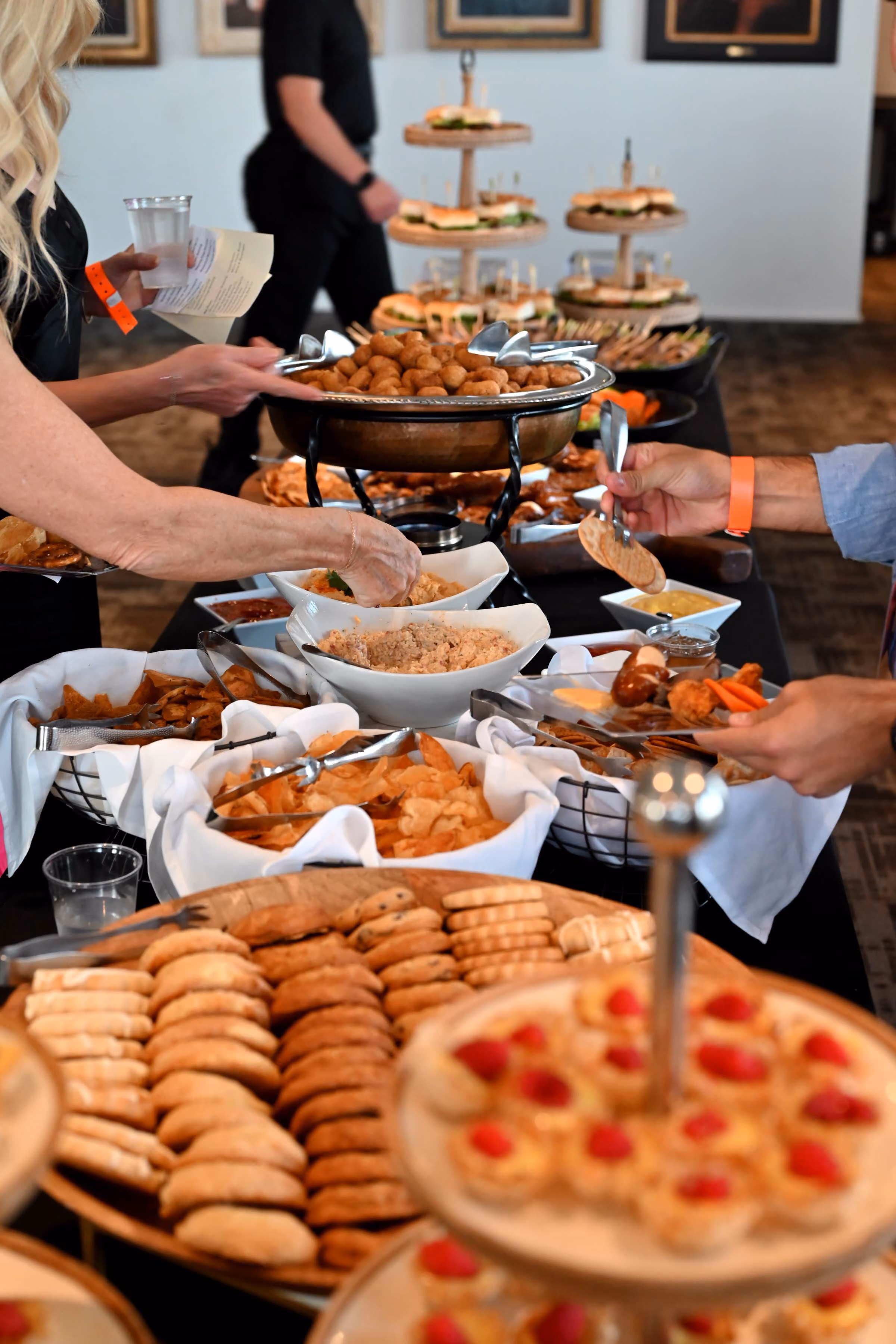Guests fill small plates at a lavish indoor buffet spread including an array of cookies, sliders, crackers, dips, chips, and other appetizers and desserts. 