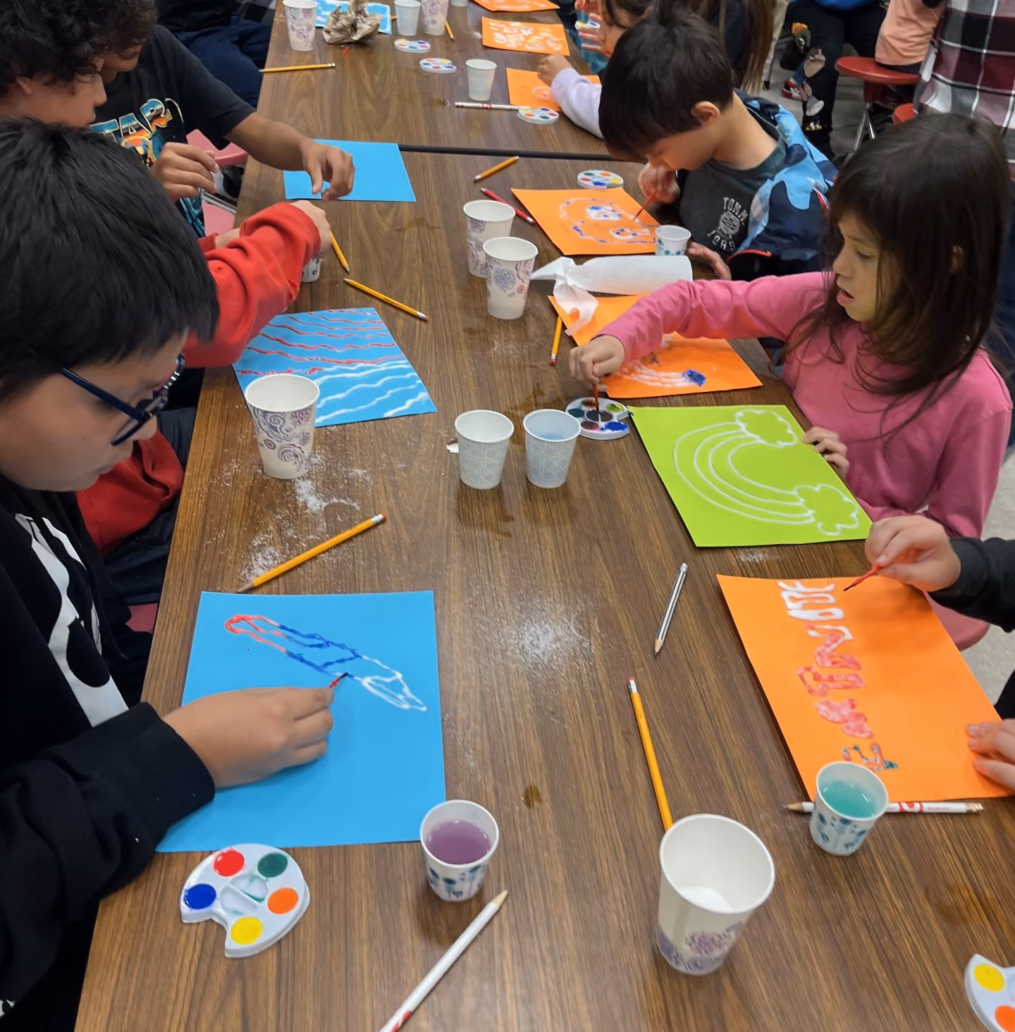 Multiple elementary school students sit around a table while creating salt rainbow art with paper, glue, salt and watercolors during an Oklahoma Originals program.