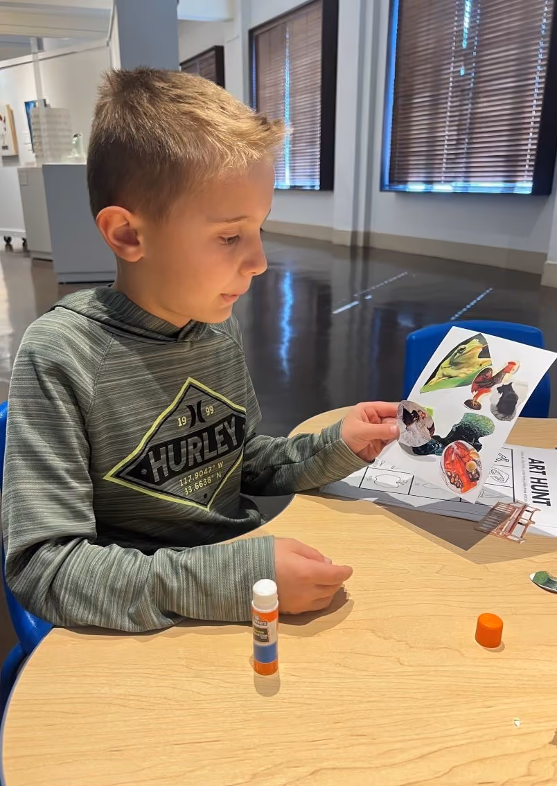 An elementary school aged boy works on a collage made out of magazine photos at a crafting station inside the art gallery.