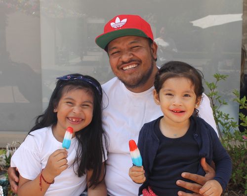 A father holds his two young girls on his lap, while both girls smile and eat red, white, and blue bomb pops in the courtyard.