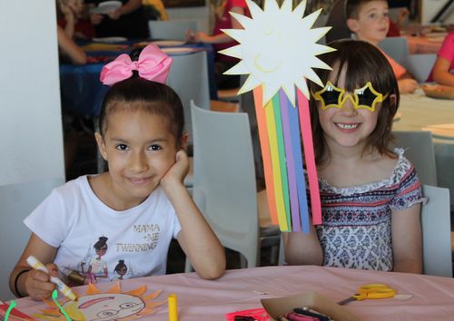 Two young girls sit at a table, crafting sun art projects. One of the girls is coloring with a marker, and the other girl is holding up her finished sun while wearing yellow. sun-shaped sunglasses.