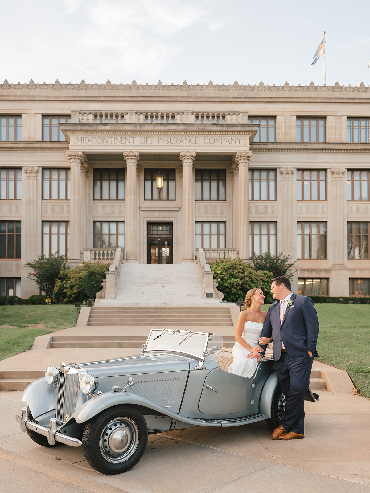 A bride and groom pose in an old car parked in front of the Oklahoma Hall of Fame building.